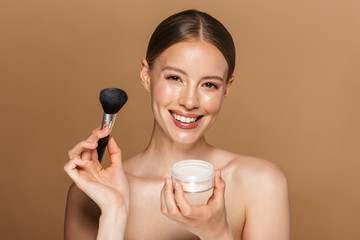 Happy beautiful young amazing woman posing isolated over brown chocolate background wall holding powder and makeup brush.