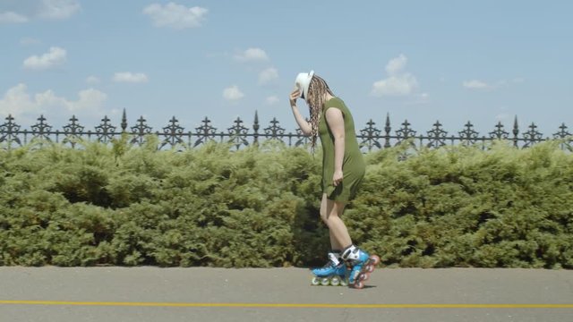Carefree joyful woman showing skill of backward crossover slide rollerblading along park alley in summer. Positive female with hat skillfully performing famous moonwalk on roller skates outdoors.