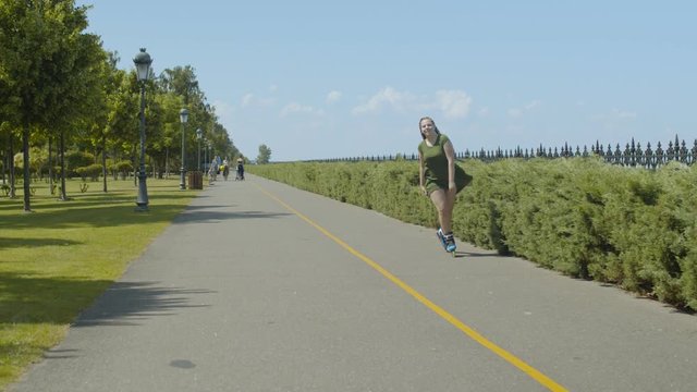 Active woman with ponytail from afro-braids rollerblading along public park walkway on windy sunny day. Positive female in loose dress enjoying summer nature and outdoor activity on roller blades.