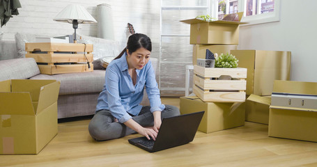 Attractive young asian chinese woman moving sitting among cardboard boxes sofa using laptop and smiling on floor. girl searching online internet website for house insurance after relocation apartment