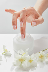 Obraz na płótnie Canvas cropped view of woman hand touching cream in jar near jasmine flowers on white surface
