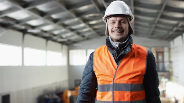 man beard and mustache in a hard hat and overalls is standing in the middle of the hangar and dancing at the camera