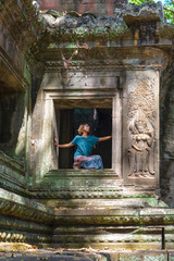 One person in Angkor Wat ruins, travel destination Cambodia. Woman in yoga position, stretching leg and raised arm, profile view, between basreliefs and stone sculptures.