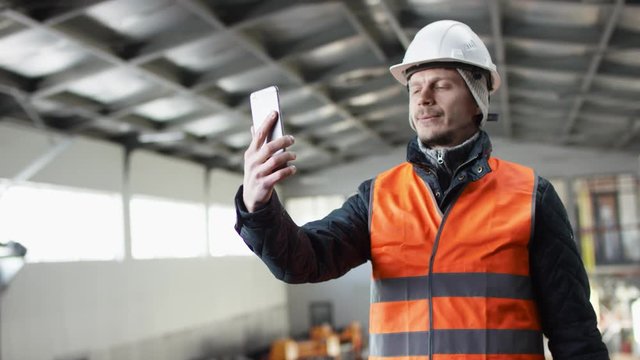 man beard and mustache in a hard hat and overalls is standing in the middle of the hangar and starting videochat by his phone at the camera