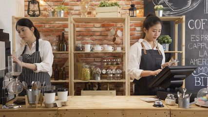 two elegant coffeehouse staff working behind counter in cafe shop. young girl waitress checking customer order with note and tablet. woman barista using machine make beverage in modern coffee store.