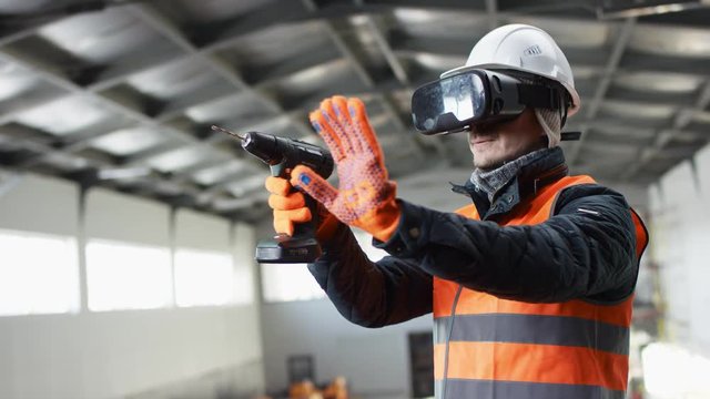 man beard and mustache in a hard hat and overalls is standing in the middle of the hangar and using drill virtual reality glasses at the camera