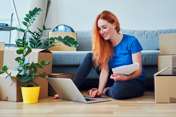 Happy young woman paying bills with laptop while sitting on floor in new apartment