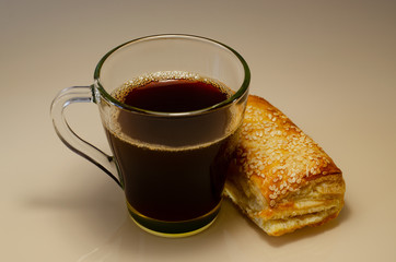 A cup of coffee in a transparent cup and a bun with sesame seeds, shot from above. Breakfast on a glass surface