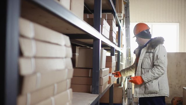 worker in a helmet and overalls stands next to the racks and remove box for right inventory with the tablet with flow camera