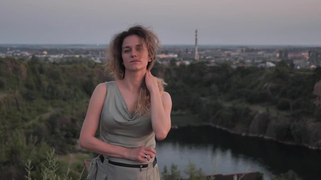 The girl looks at the camera at sunset on the mountain against the town. She stands in her dress and straightens her curly hair . View from the hill to the city.