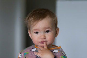 Closeup of cute fair sleepy eyed toddler girl eating yogurt and putting a finger in her mouth while sitting in her high chair with a bib