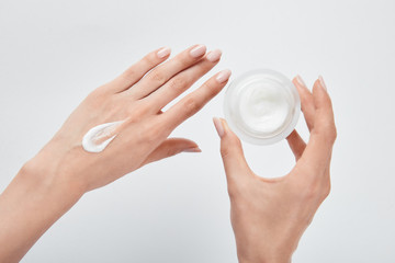 cropped view of woman applying cream on skin and holding jar isolated on white