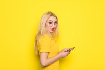 Portrait of blonde girl having smart phone in hands using wi-fi 4G internet checking email chatting with friends searching contact isolated on yellow background