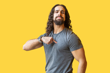 Portrait of proud bearded young man with long curly hair in grey tshirt standing, pointing himself...