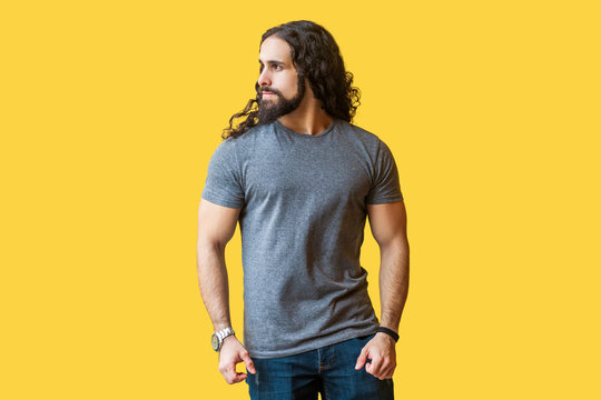 Portrait Of Brutal Handsome Bearded Young Man Model With Long Curly Hair In Grey Tshirt Standing And Looking Away With Serious Proud Face. Indoor Studio Shot Isolated On Yellow Background.