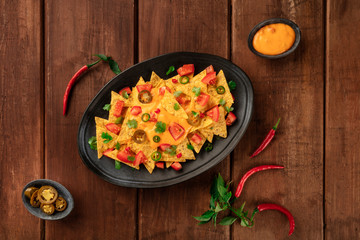 Nachos, Mexican tortilla chips, shot from the top with a cheese sauce, chili and jalapeno peppers, tomatoes. and cilantro, on a dark rustic wooden background