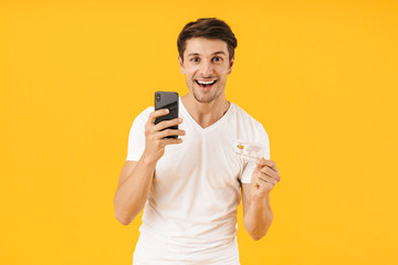 Photo of brunette man in basic t-shirt smiling at camera while holding smartphone and credit card