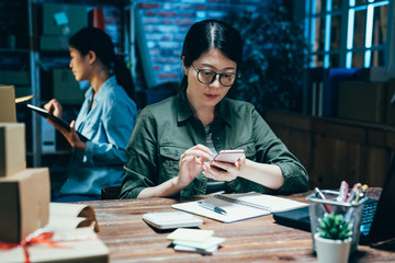 Fototapeta premium individual e commerce business at house studio. young hard working girl freelancer check orders and packing goods for delivery on mobile phone. employee colleague using tablet out time work night