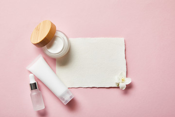 top view of open jar with cream, cosmetic glass bottle, cream tube, piece of paper and jasmine flower on pink