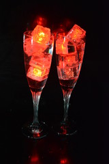 Two unusual glasses of champagne with a drink and bright red ice chips. Plastic ice cubes of ice illuminating the drink in each glass on a black background, an impressive photo and the feast of St. Va