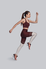 Fototapeta na wymiar Always in good shape. Full length of attractive young woman in sports clothing jumping while exercising against grey background