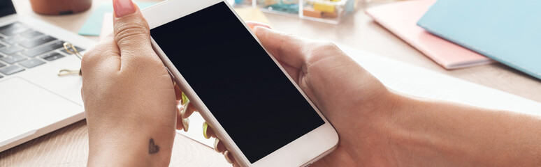 cropped view of woman holding smartphone in hands over wooden table with laptop and stationery