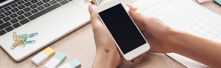 cropped view of woman holding smartphone in hands over wooden table with laptop, planners and stationery