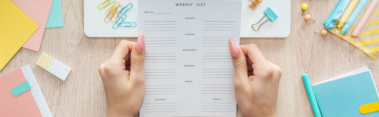 cropped view of woman holding weekly list over wooden table with laptop and stationery