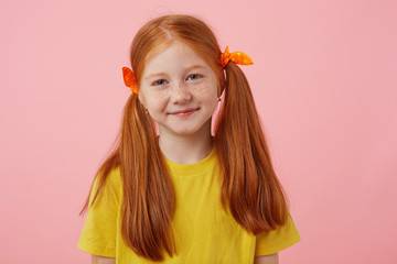Close up of happy petite freckles red-haired girl with two tails, smiling and looks cute, wears in yellow t-shirt, stands over pink background.