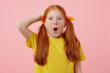 Portrait of happy amazed petite freckles red-haired girl with two tails, wears in yellow t-shirt, stands over pink background with wide open mouth.
