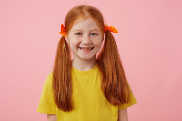 Portrait of happy petite freckles red-haired girl with two tails, broadly smiling and looks cute, wears in yellow t-shirt, stands over pink background.