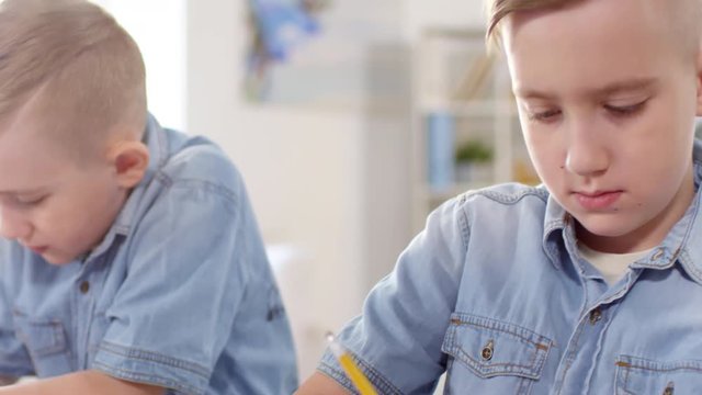 Chest-up panning tilting shot of 10-year-old identical twin brothers, dressed in denim shirts, sitting side by side and drawing with pencils in copybooks