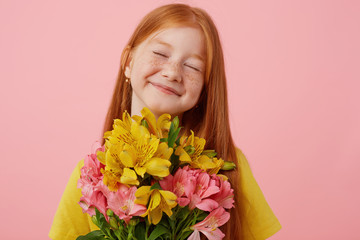 Fototapeta premium Portrait petite freckles red-haired girl with two tails, broadly smiling and looks cute, with closed eyes, holds bouquet, wears in yellow t-shirt, stands over pink background.