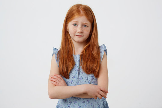 Portrait of little unhappy freckles red-haired girl with two tails, malcontent looks at the camera, wears in blue dress, stands with crossed arms over white background.