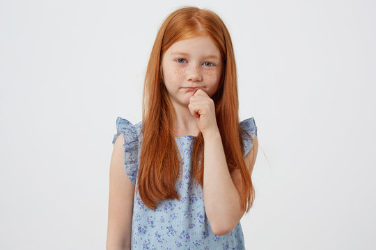 Portrait of little unhappy freckles red-haired girl, sadly looks at the camera, wears in blue dress, stands over white background.
