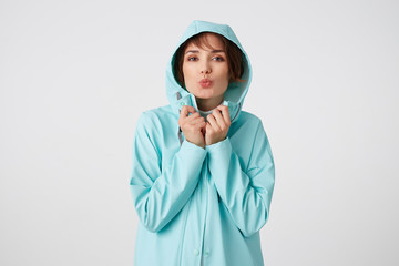 Portrait of positive young nice lady in blue rain coat, with a hood on his head, looks at the camera with happy expressions, send kiss at the camera, stands over white wall.