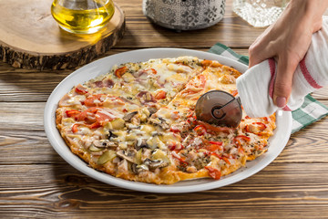 Closeup hand of female cutting pizza with tomatoes, ham and mushroom on wooden table