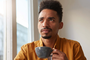 Portrait of young beauty African American thinking boy thoughtfully looks up, drinks aromatic coffee from a gray cup , trying to remember something