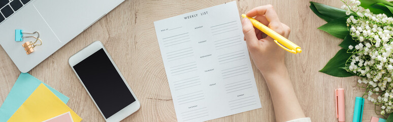 cropped view of woman sitting behind wooden table with smartphone and laptop, witting in weekly list