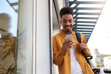 Young cheerful African American guy in yellow shirt, walking down the street and holds telephone, got a message with a funny kitten, looks happy and broadly smiling. © timtimphoto