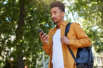 Fototapeta premium Young frowning African American man in yellow shirt, chatting on the phone with his friends and waiting him at the park, looks up with resentful expression, his friend late.