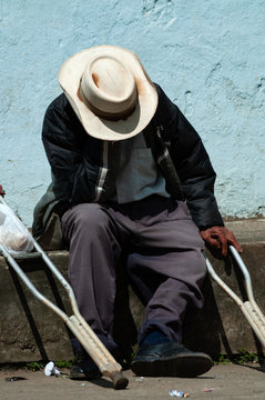 Hombre con sombrero y muletas sentado en la acera de la calle de un pueblo