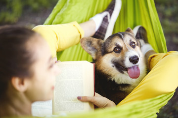 Smiling young woman reading a book and lying in green hammock with funny dog Welsh Corgi in a park outdoors. Closeup portrait of doggy. Concept friendship with dog and human, cute moments, relaxing