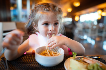 Litle girl is eating pasta in the restaurant.