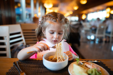 Litle girl is eating pasta in the restaurant.