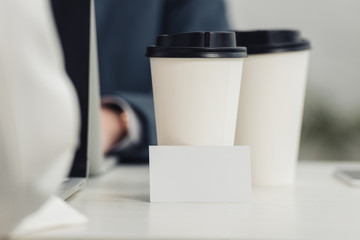 selective focus of blank business card and disposable cups on table near businesspeople using laptops