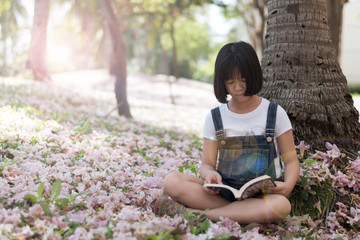 Adorable Asia female kid reading literacy in park. Child reader read young adult novel book in garden park with pink flowers o ground.