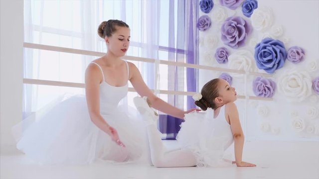 Little child girl ballerina in white tutu makes stretching exercise in ballet class. She is stretching in frog pose with her teacher help on lesson. Classical ballet training.
