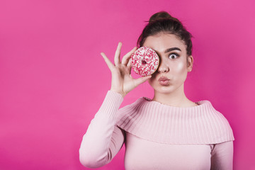 Surprised young woman pouting her lips covering her eyes with donut on pink backdrop