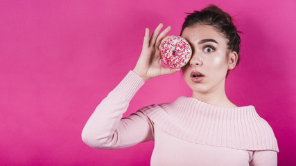 Afraid young woman holding donut over her eyes against pink backdrop
